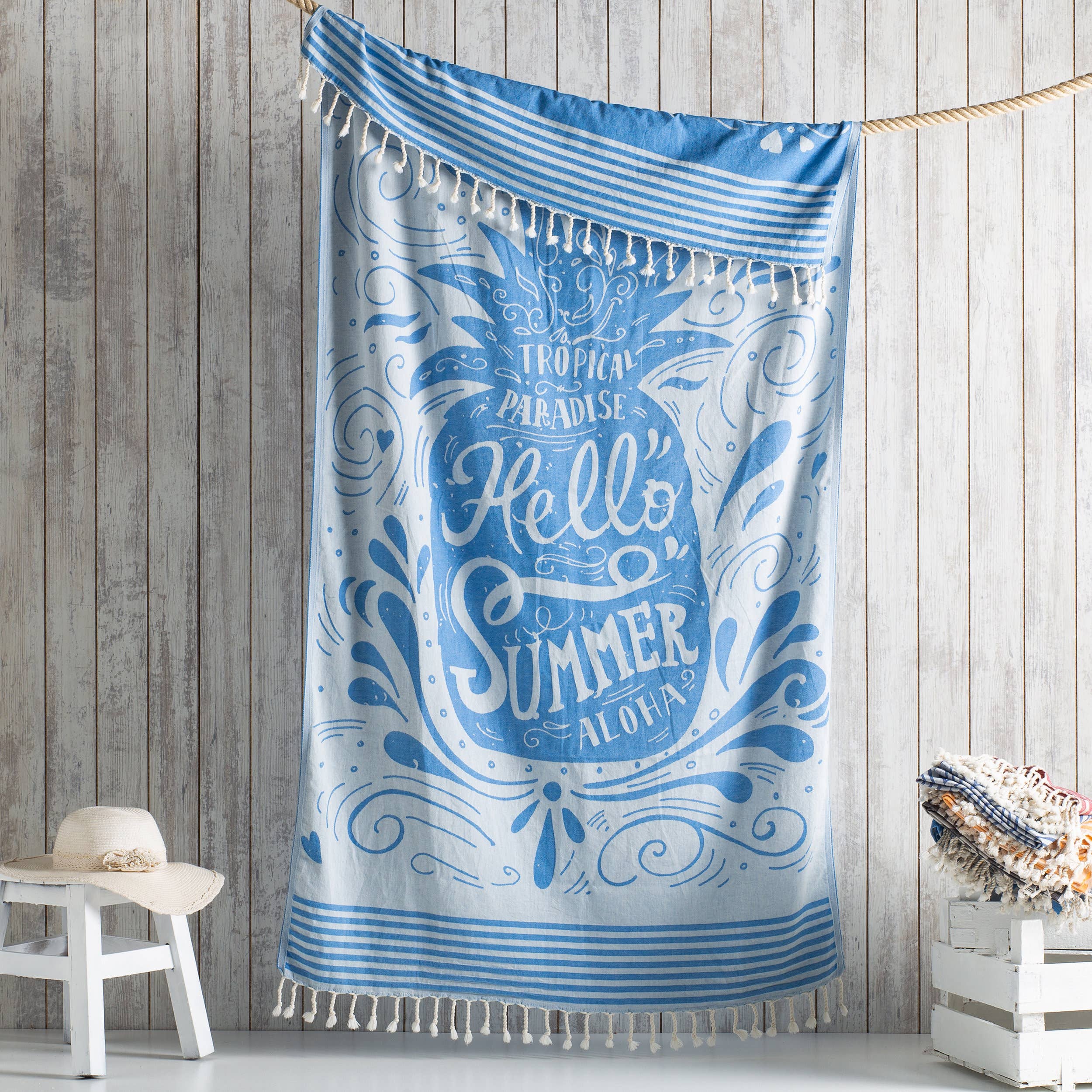 A blue and white Turkish cotton beach towel with a fouta design, hanging on a wooden wall, featuring text 'Hello Summer' and decorative elements.