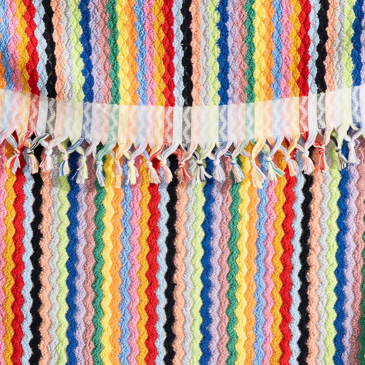 A close-up of a colorful striped woven textile with vertical zigzag bands in red, orange, yellow, green, blue, pink, black, and white, finished with white lace trim and tassel fringe.