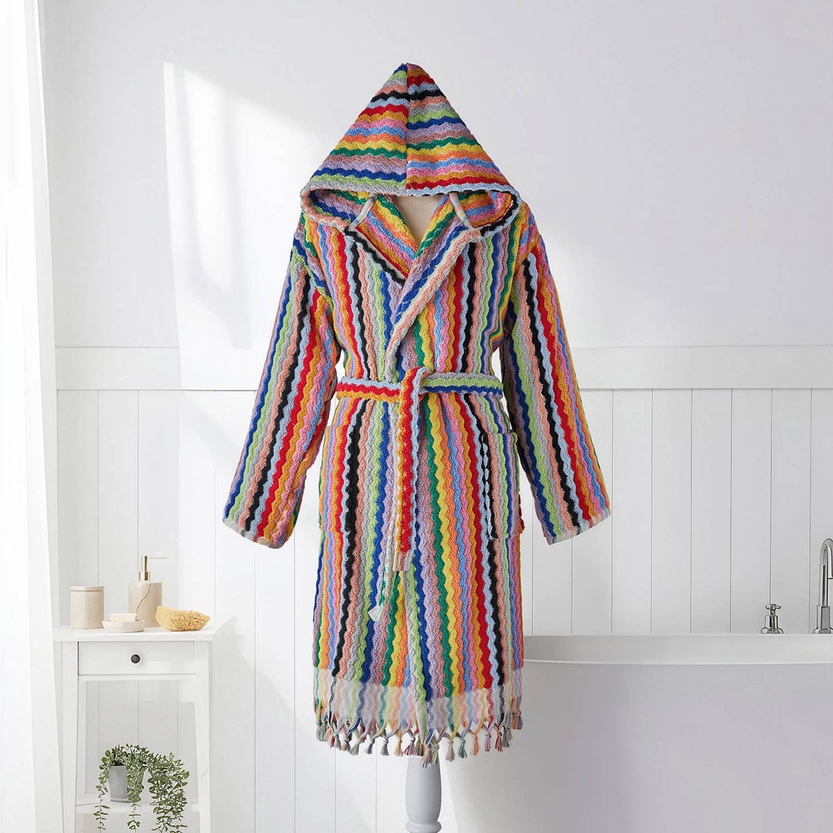 A multicolor striped knit hooded robe with a tie belt and tassel fringe, displayed on a mannequin in a bright white bathroom setting.