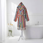 A multicolor striped hooded bathrobe with tassels hanging from the hem, displayed on a white tripod stand in a bright, modern bathroom setting with a bathtub and white wall paneling.