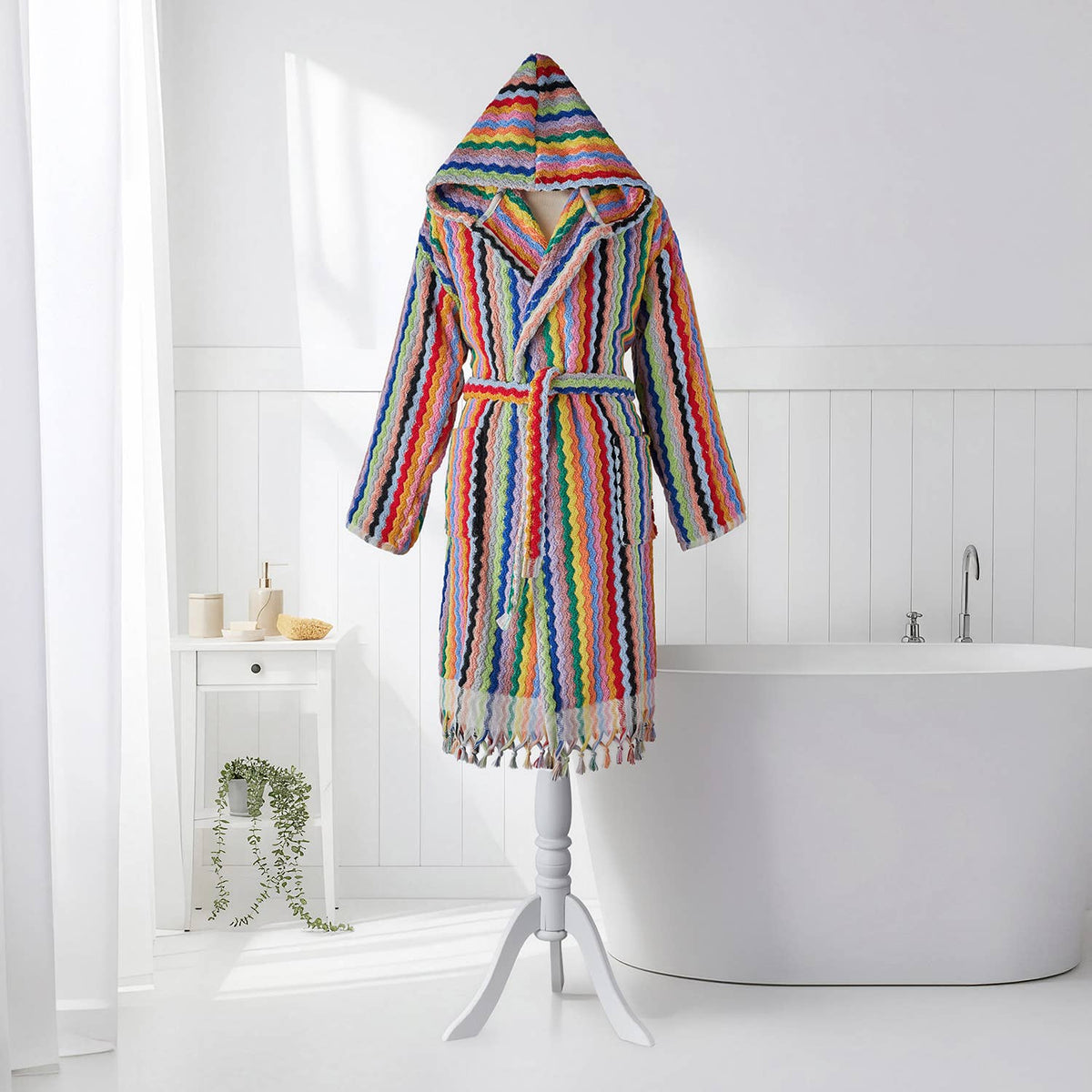 A multicolor striped hooded bathrobe with tassels hanging from the hem, displayed on a white tripod stand in a bright, modern bathroom setting with a bathtub and white wall paneling.