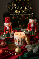 A festive holiday scene with two nutcracker figurines flanking a lit candle in a glass jar. The candle is decorated with colorful nutcracker illustrations and sits on red draped fabric with pine branches, ornaments, and warm string lights.