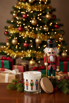 A lit white candle with a glass base and a lid, printed with colorful nutcracker figures, sits on a wooden surface in front of a decorated Christmas tree and gift boxes. A wooden nutcracker figurine stands beside the candle.