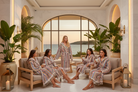 Four women wearing matching multicolor striped bathrobes relax in a luxurious spa lounge with a sea view background.