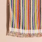 A close-up of a colorful striped textile with tassel fringe, shown hanging against a warm beige background. The fabric features vertical wavy stripes in many pastel and bright colors, including pink, yellow, blue, green, orange, red, and black.