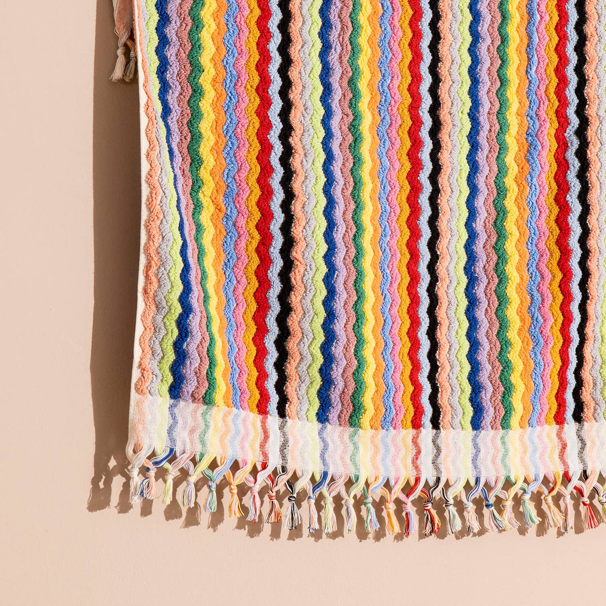 A close-up of a colorful striped textile with tassel fringe, shown hanging against a warm beige background. The fabric features vertical wavy stripes in many pastel and bright colors, including pink, yellow, blue, green, orange, red, and black.
