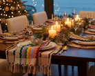 A beautifully set dining table for six with white plates, gold cutlery, colorful striped knit placemats, and lit candles, set against a window view of the ocean at dusk with a decorated Christmas tree in the background.