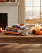 A stack of folded multicolored knit blankets with fringe is displayed on a wooden table in a cozy autumn living room setting, with pumpkins, candles, a fireplace, and a window in the background. The text 'Give Thanks in Style' is visible in the bottom right corner.