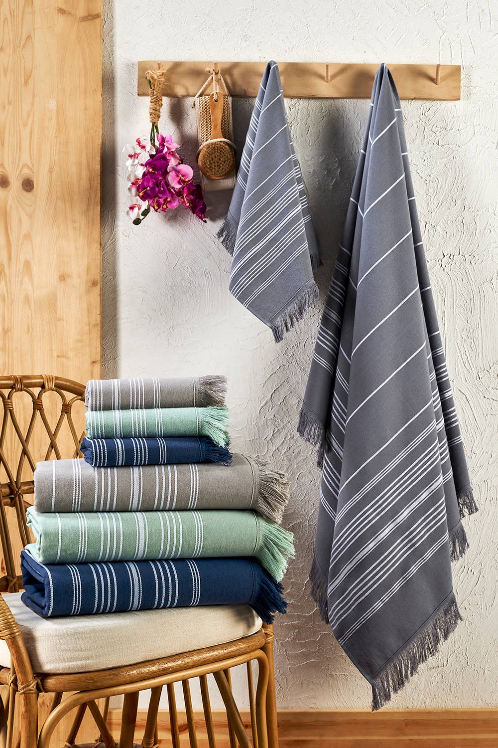 A set of woven towels and throws in gray, navy, and mint green with white stripes and fringed edges, displayed hanging on a wooden rack and stacked on a wicker chair against a textured wall.