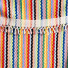 A close-up of a colorful striped woven textile with vertical zigzag bands in red, orange, yellow, green, blue, pink, black, and white, finished with white lace trim and tassel fringe.