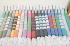 A row of colorful folded woven towels with white fringe, each labeled with a color name above it, including mint green, gray, dark gray, light gray, yellow, orange, pink, lavender, black, navy, blue, and green.