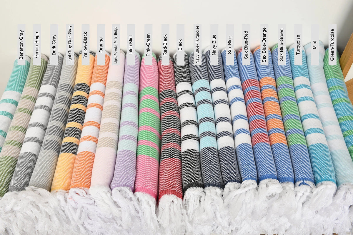 A row of colorful folded woven towels with white fringe, each labeled with a color name above it, including mint green, gray, dark gray, light gray, yellow, orange, pink, lavender, black, navy, blue, and green.