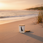 A white glass jar candle with a light wood lid sits on a sandy beach at sunset, with the matching lid placed beside it. The background shows ocean waves, dunes, and warm golden light.