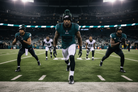A group of Philadelphia Eagles football players run onto the field during a game, with the central player in a dark teal Eagles jersey and white pants running forward under stadium lights.