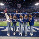 Five women in matching New York Giants apparel pose on a football field under falling confetti. They wear blue and white Giants beanies, sweatshirts, and jeans, with one person holding a football and another holding a foam finger.