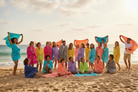 A group of people wrapped in colorful towels and standing or sitting on a sandy beach, each holding up a different towel color including turquoise, pink, orange, yellow, purple, and blue.