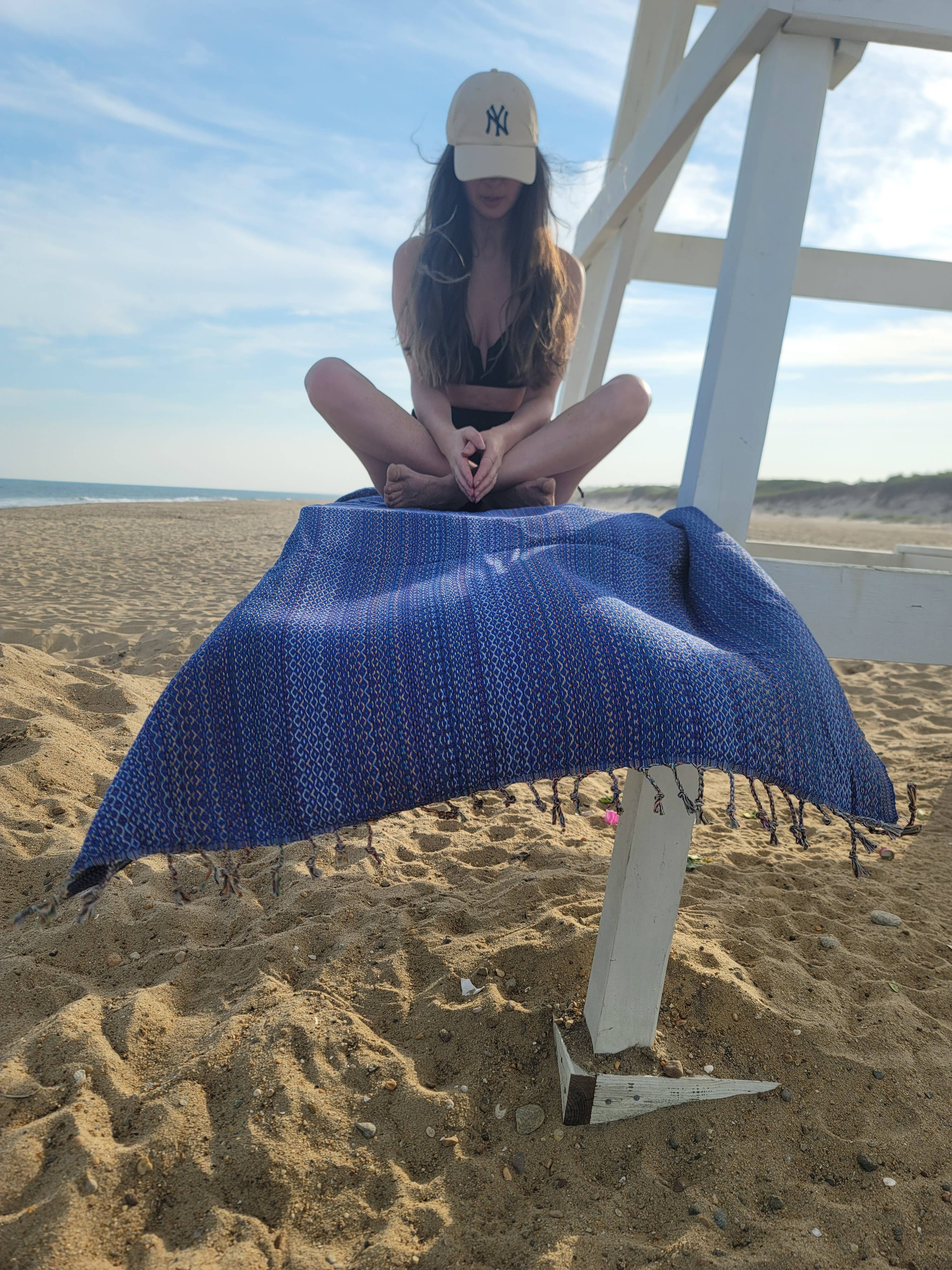 A stack of folded handwoven beach towels in various colors, including blue, green, red, and brown, with a small potted plant and sunglasses beside them.