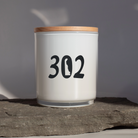 A white glass jar candle with a light wood lid sits on a textured gray stone slab against a neutral gray background. The front of the candle is printed with the black numbers '302'.