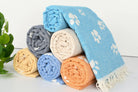A stack of rolled towels in multiple colors, including light blue, gray, cream, yellow, and orange, with white paw print patterns and fringed edges, set against a clean white background with a small green plant on the left.