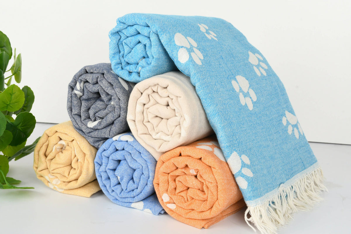 A stack of rolled towels in multiple colors, including light blue, gray, cream, yellow, and orange, with white paw print patterns and fringed edges, set against a clean white background with a small green plant on the left.