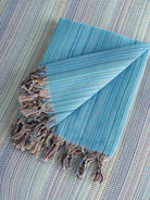 A stack of folded handwoven beach towels in various colors, including blue, green, red, and brown, with a small potted plant and sunglasses beside them.