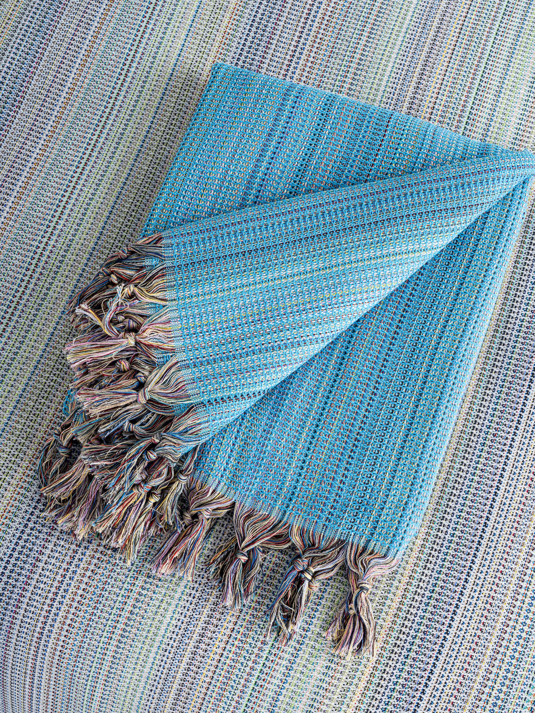 A stack of folded handwoven beach towels in various colors, including blue, green, red, and brown, with a small potted plant and sunglasses beside them.