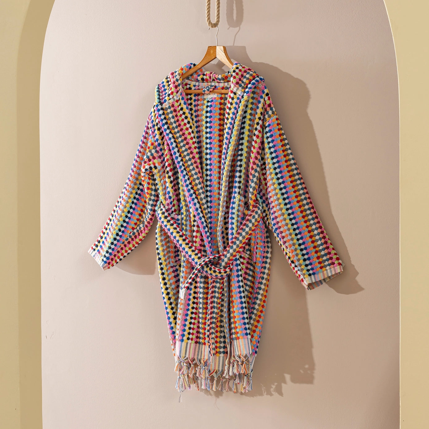 A multicolor woven robe with a tie waist and tassel fringe, hanging on a wooden hanger against a beige arched wall.