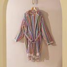 A multicolor woven robe with a tie waist and tassel fringe, hanging on a wooden hanger against a beige arched wall.