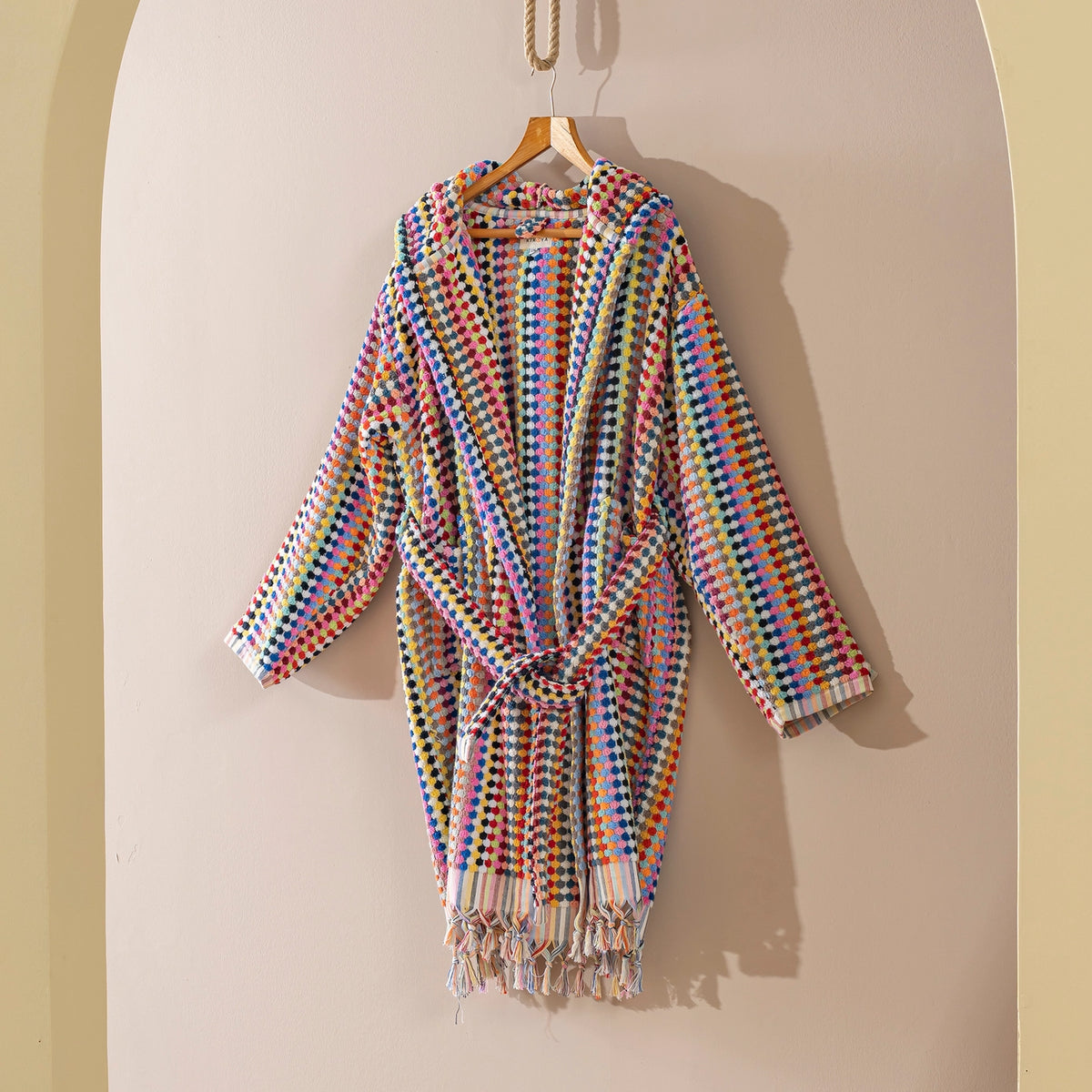 A multicolor woven robe with a tie waist and tassel fringe, hanging on a wooden hanger against a beige arched wall.