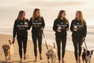 Four women walking dogs on a beach at sunset, wearing matching black hoodies with the text 'My Therapist Barks' in white and orange lettering.