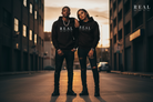 Two models standing on a city street at sunset wearing matching black hoodies with 'REAL' printed in white across the chest, paired with distressed black jeans and black boots. The brand logo 'REAL' is also visible in the top right corner.