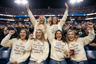 Five women cheering in a crowded sports arena, wearing matching cream-colored hoodies with colorful text that reads 'DO WHATEVER THE FUCK MAKES YOU HAPPY'.