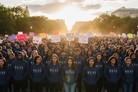 A large crowd of people standing outdoors at sunset, many wearing matching navy blue hoodies with the word 'REAL' printed in white across the chest. Several people hold protest-style signs, including one that reads 'LOVE WINS.