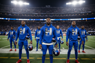 A group of football players standing on a stadium field at night, wearing matching royal blue hoodies and pants with a large white 'H' and 'HOCKINGTON' text on the front, each holding a helmet.