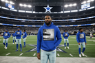 A man stands on a Dallas Cowboys football field wearing a royal blue hoodie with a black-and-white graphic of lips and the word 'KUSHHHHH' on the front, paired with light blue pants. Other Dallas Cowboys players in matching hoodies are visible in the background inside the stadium.