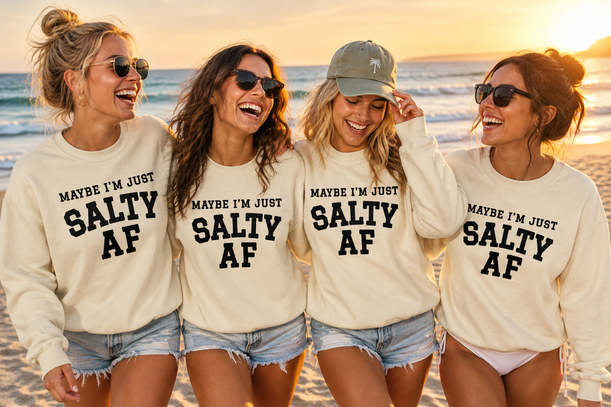 Four women standing together on a beach at sunset, all wearing matching cream sweatshirts with bold black text that reads 'MAYBE I'M JUST SALTY AF' and denim shorts.