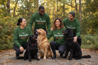 Four people wearing matching green crewneck sweatshirts with the text 'My Therapist Barks' pose outdoors with three dogs on a stone path in a wooded area.