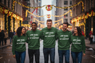 Five people standing together on a city street decorated with festive Christmas lights and snowfall, wearing matching green crewneck sweatshirts with the slogan 'MY GOD IS MY ENEMIES' printed in white across the front.