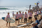 Surfers in pink shirts with surfboards on a beach with waves and flags in the background