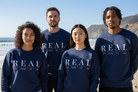 Four people standing outdoors on a beach wearing matching navy blue sweatshirts with the word 'REAL' printed in large white letters across the chest.
