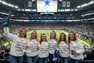 Five women standing together in a crowded football stadium, wearing matching white long-sleeve shirts with the slogan 'DO WHAT THE FUCK MAKES YOU HAPPY' printed in red, blue, and purple text. One woman on the left holds a blue foam finger.